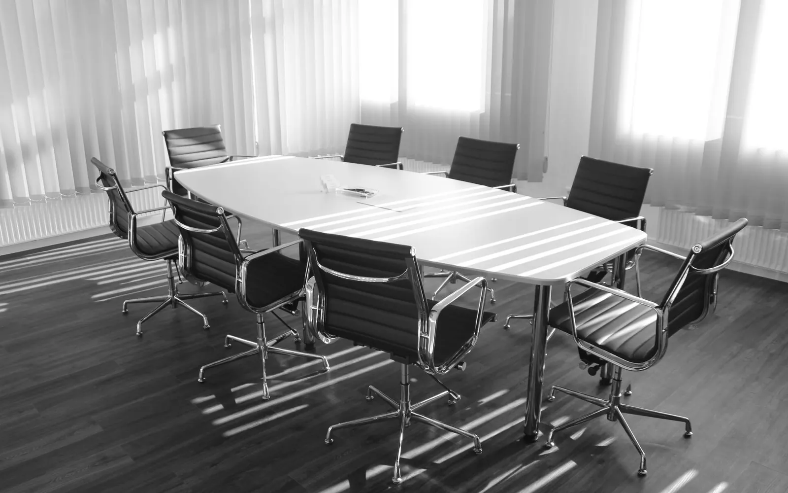 An empty boardroom table with eight chairs in low sunlight through vertical blinds